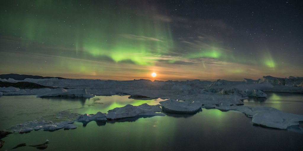 A rising moon helps the northern lights to illuminate icebergs trapped within the Ilulissat Icefjord, a UNESCO World Heritage Site since 2004. The icebergs are calved from the Sermeq Kujalleq glacier located roughly 60 miles up fjord to the east. The town of Ilulissat lies 2 miles to the north of the Icefjord's mouth in western Greenland. September 2016